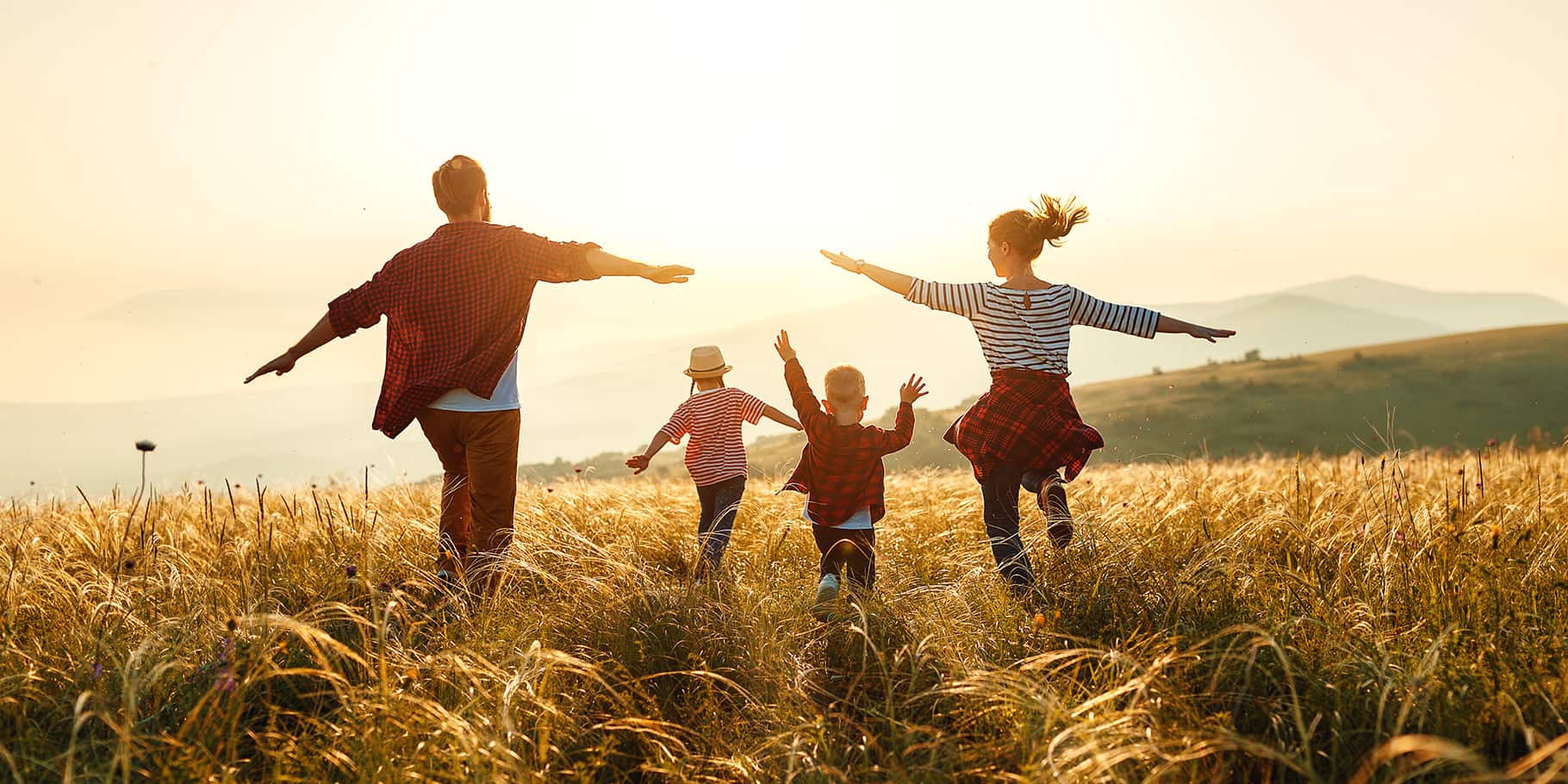 A young family of four running through an open wheat field with outstretched arms.
