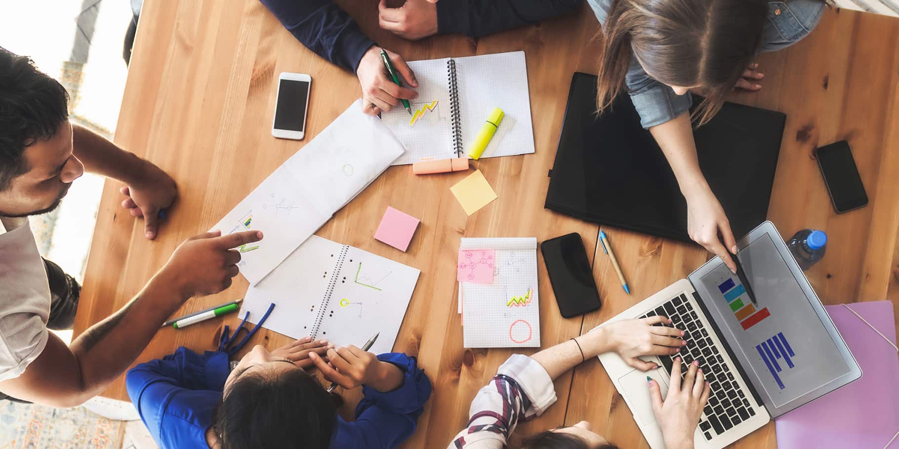 A top-view of 5 young professionals sitting at a table working on financial charts.